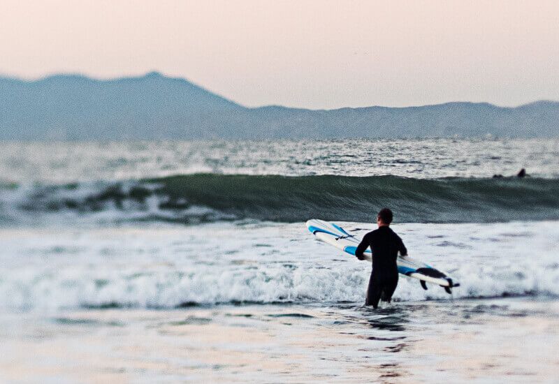 Hombre con tabla de surf entrando en el mar, imagen asociada a un estilo de vida activo y consciente.